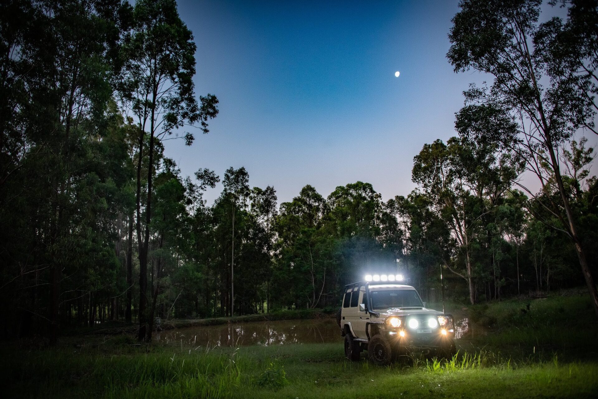 Toyota LandCruiser 76 Series off-road at dusk with headlights and roof lights illuminating in front of a forest and river.