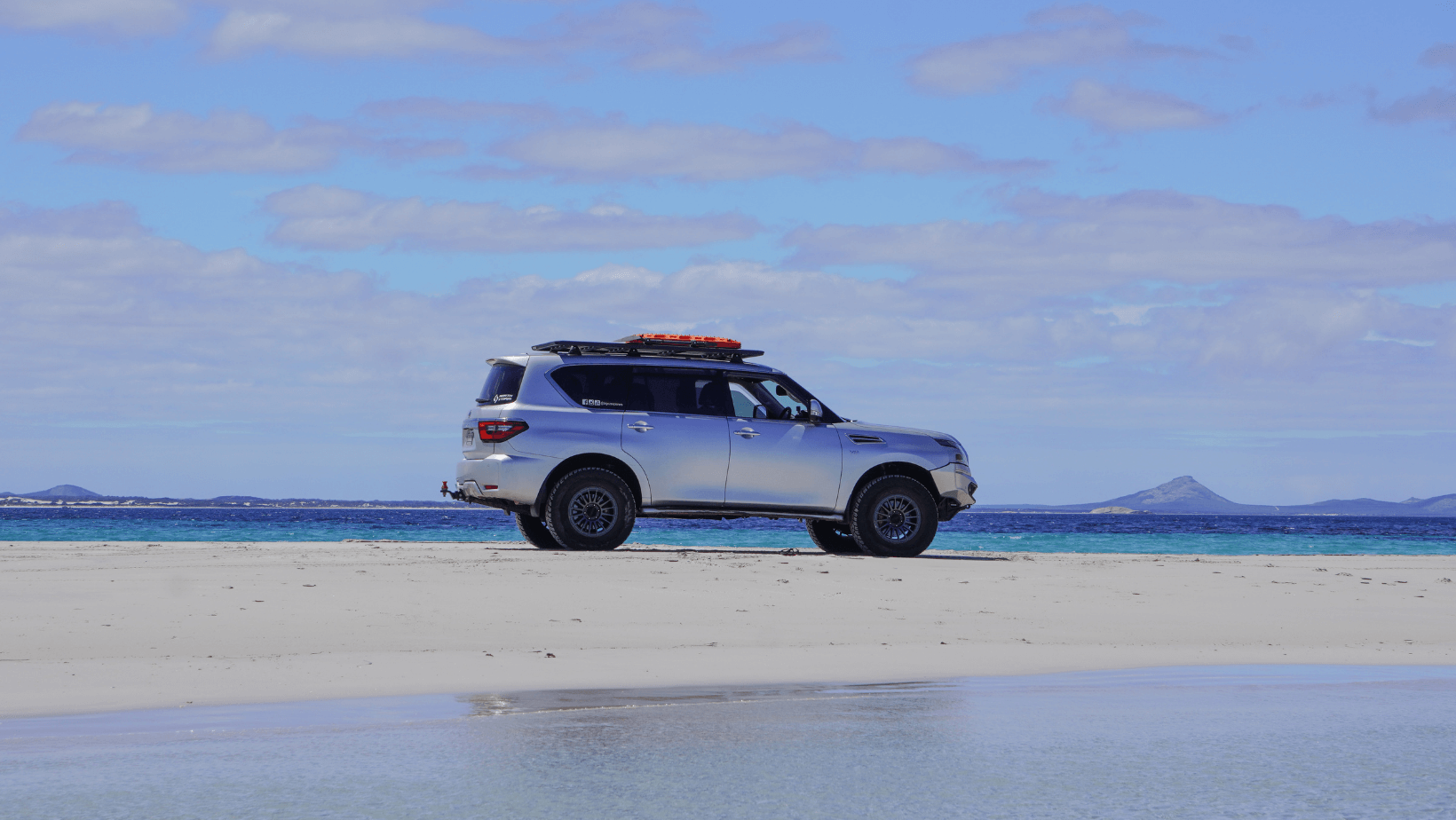 Silver 4WD parked on white sand beach near turquoise ocean with blue sky and mountains in the distance.
