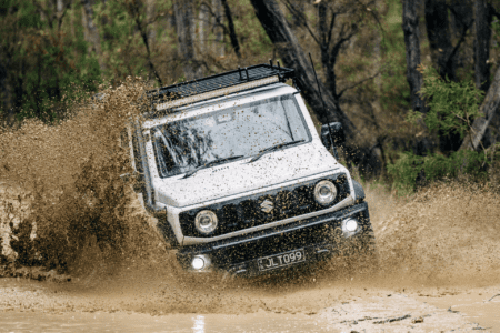 White Suzuki Jimny driving through muddy off-road terrain, splashing water.