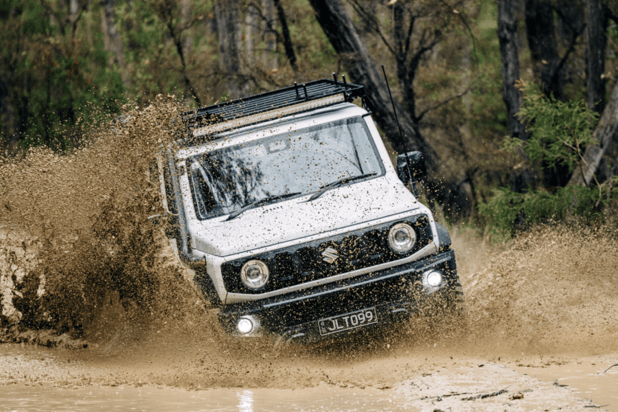 White Suzuki Jimny driving through muddy off-road terrain, splashing water.